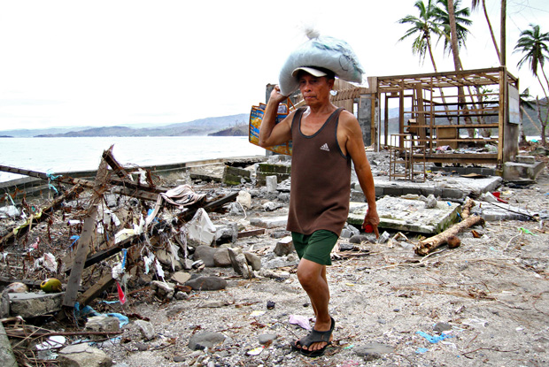 A resident carrying relief goods walks past houses damaged by Typhoon Pablo at Ugpason Beach, Barangay San Roque, in the town of Lingig, Surigao del Sur on Christmas Day. MindaNews photo by Erwin Mascarinas